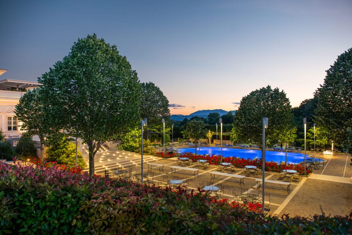 Esterno serale di Villa Orsini con piscina illuminata, terrazza con tavoli in ferro battuto e giardini fioriti sullo sfondo collinare