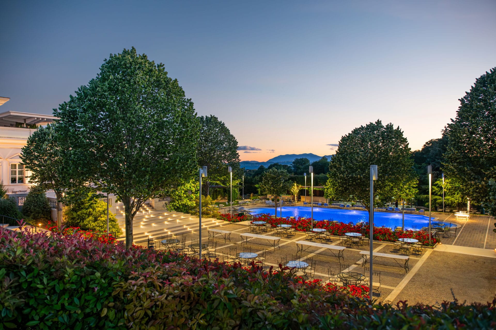 Esterno serale di Villa Orsini con piscina illuminata, terrazza con tavoli in ferro battuto e giardini fioriti sullo sfondo collinare