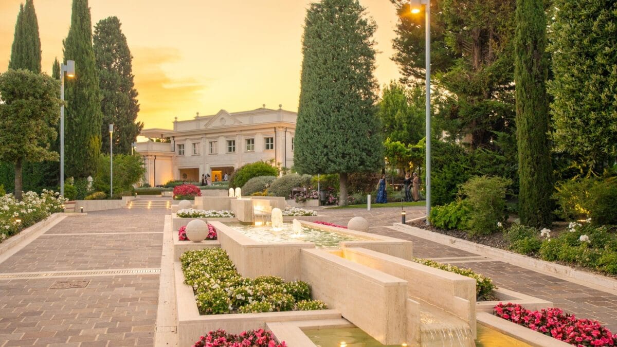 La facciata di Villa Orsini al tramonto, circondata da alberi con fontane e fiori in primo piano